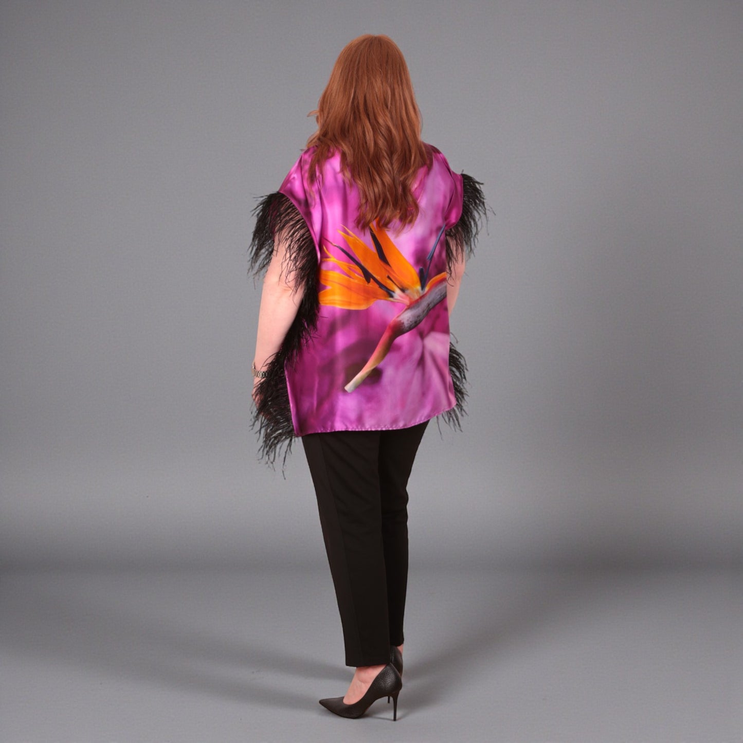 Women wearing a pink purple top with bird of paradise flower design and black feather trim with black trousers on a plain background. Photographed from the back