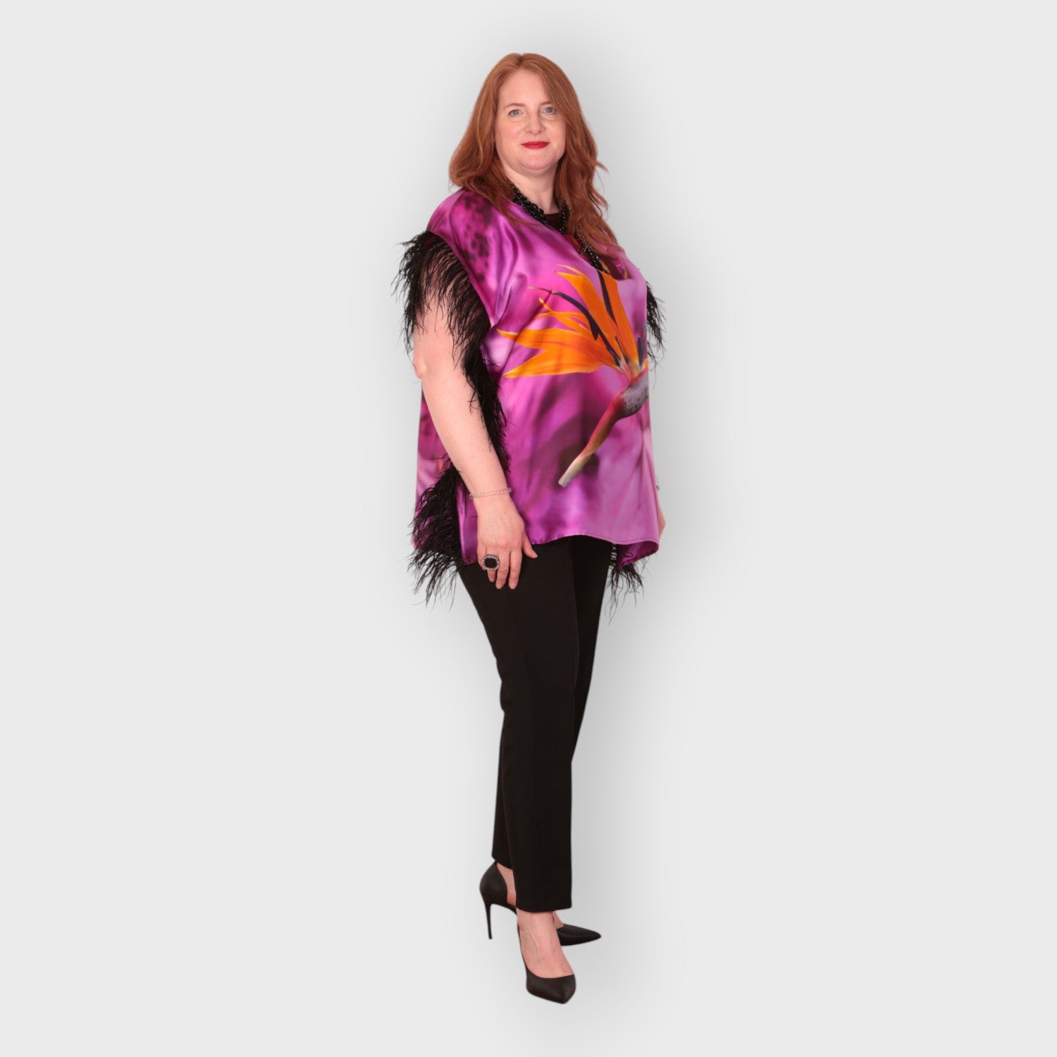 Women wearing a pink purple top with bird of paradise flower design and  black feather trim with black trousers on a white background
