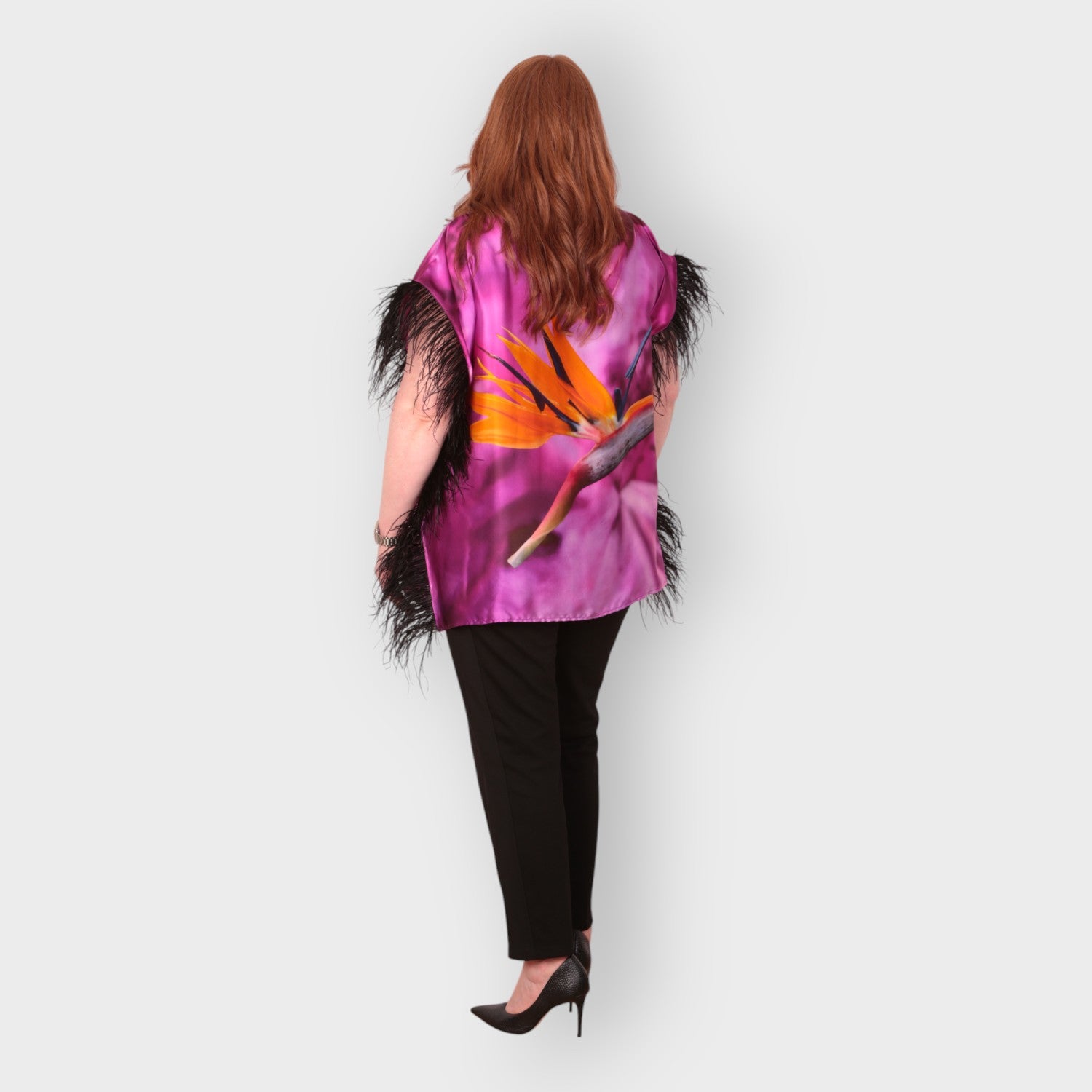 Women wearing a pink purple top with bird of paradise flower design and  black feather trim with black trousers on a white background. Photographed from the back