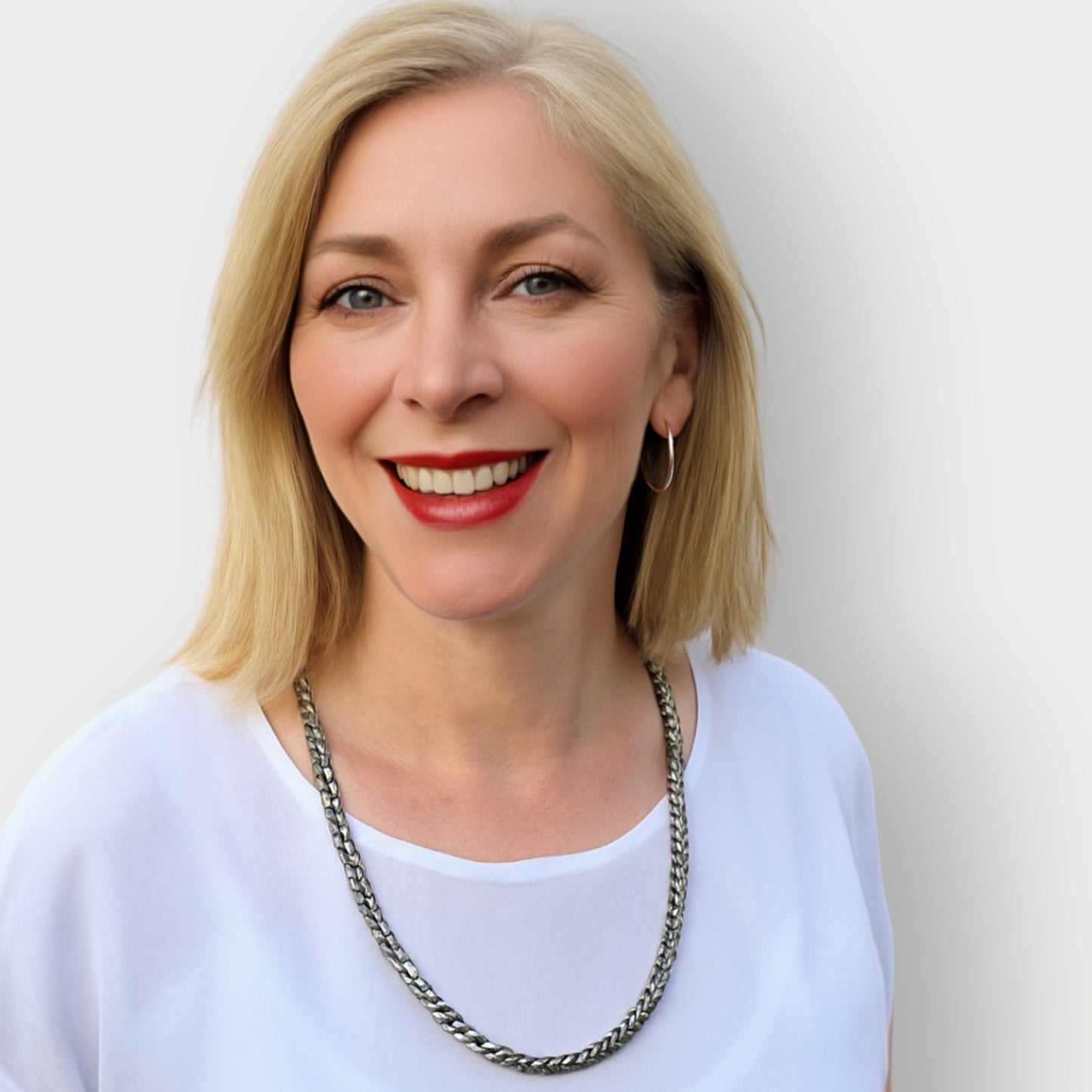 Woman with blonde hair and red lipstick wearing a white top and silver necklace against a plain background