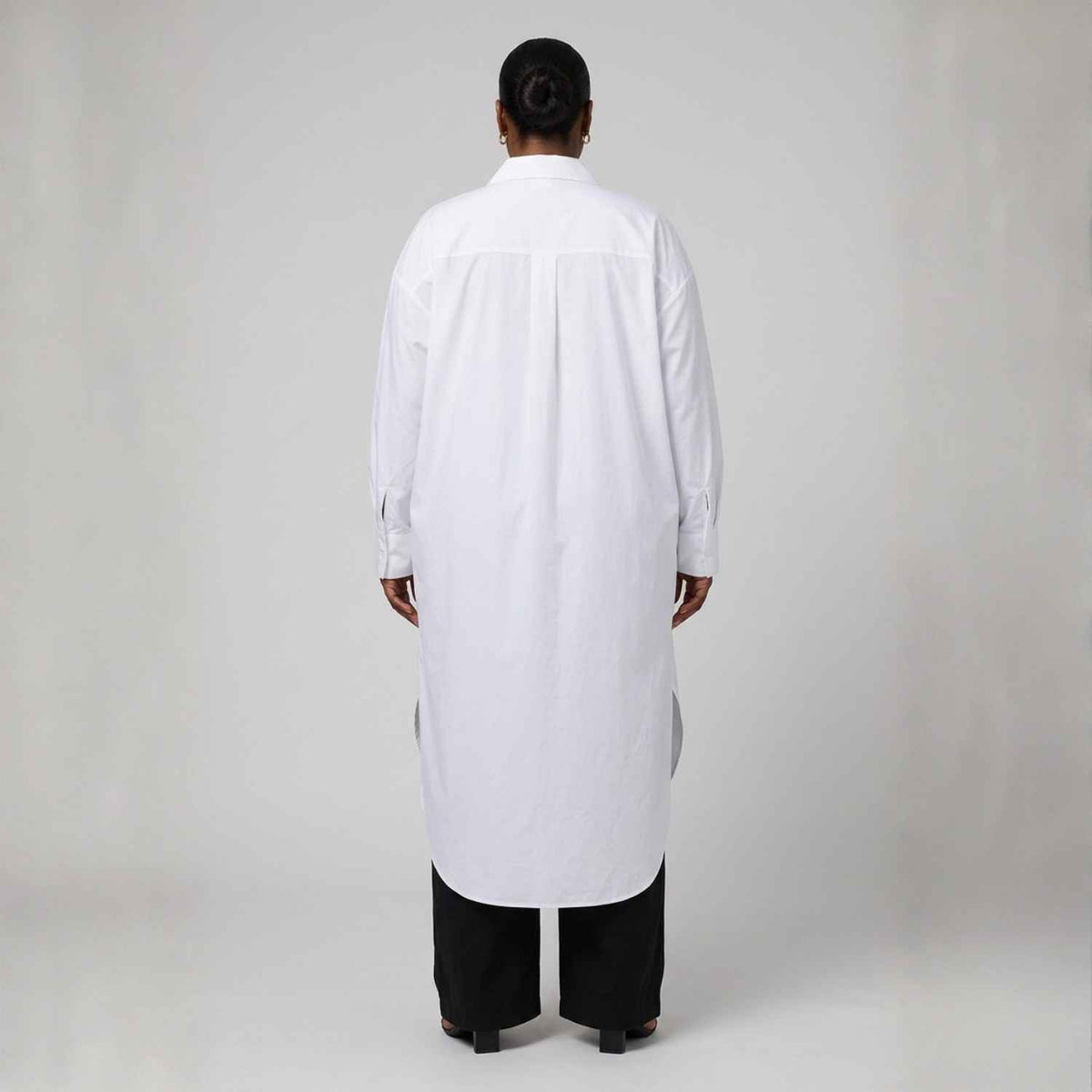A person viewed from the back wearing a long white button-up shirt or tunic over black pants, standing against a plain light gray studio background.