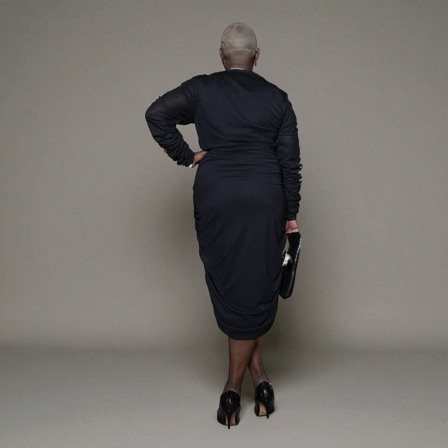 Plus-size woman wearing a black Boudicca Ruched Bodycon Midi Dress, styled with black heels. Plain background.