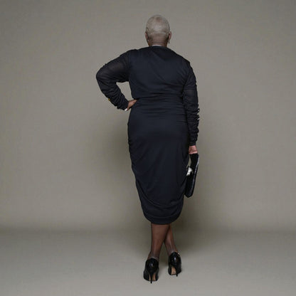 Plus-size woman wearing a black Boudicca Ruched Bodycon Midi Dress, styled with black heels. Plain background.