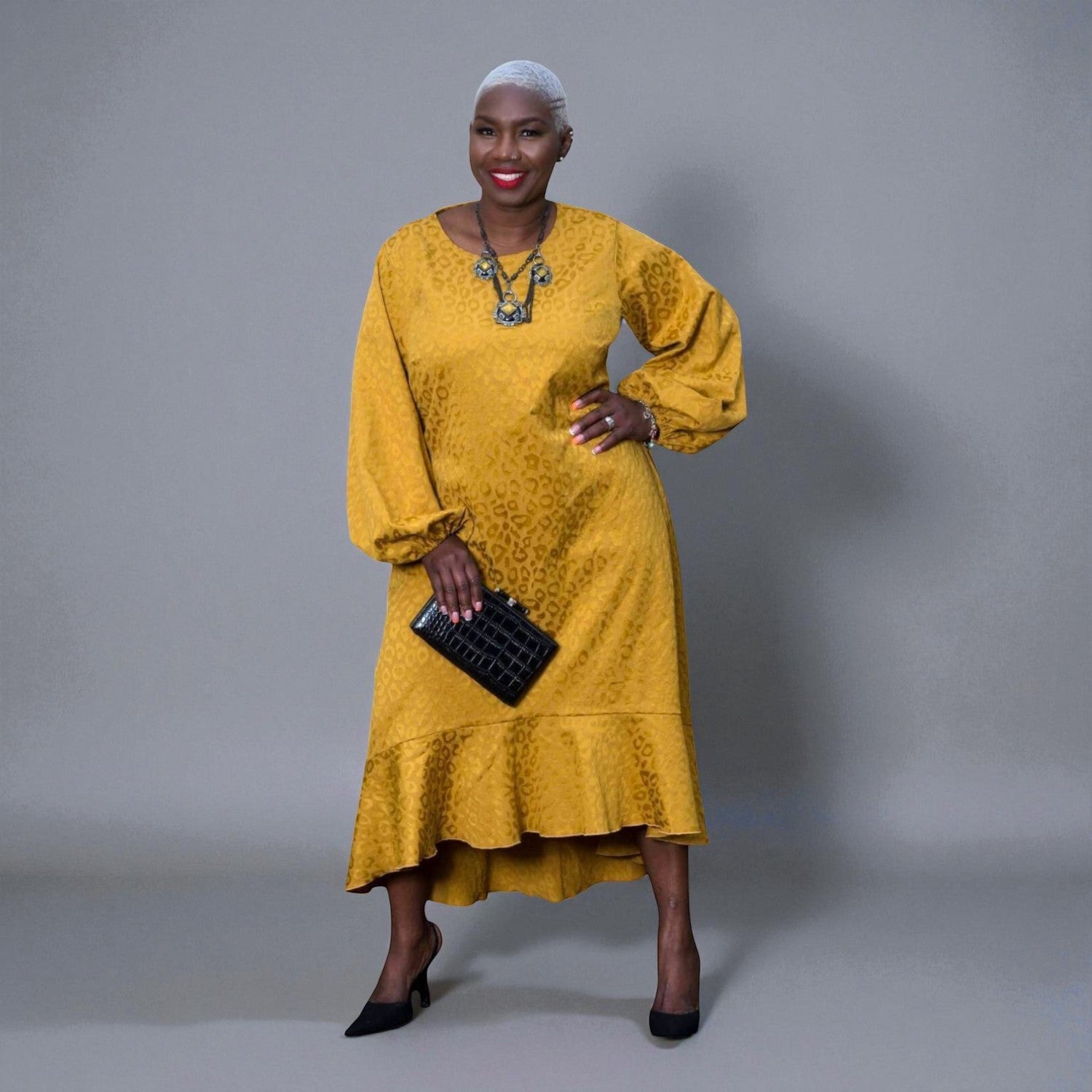 Women's Plus Size Gold Aphrodite Leopard Jacquard Dress styled with a yellow stone necklace, black clutch, and black heels for an elegant look. Gray Background.
