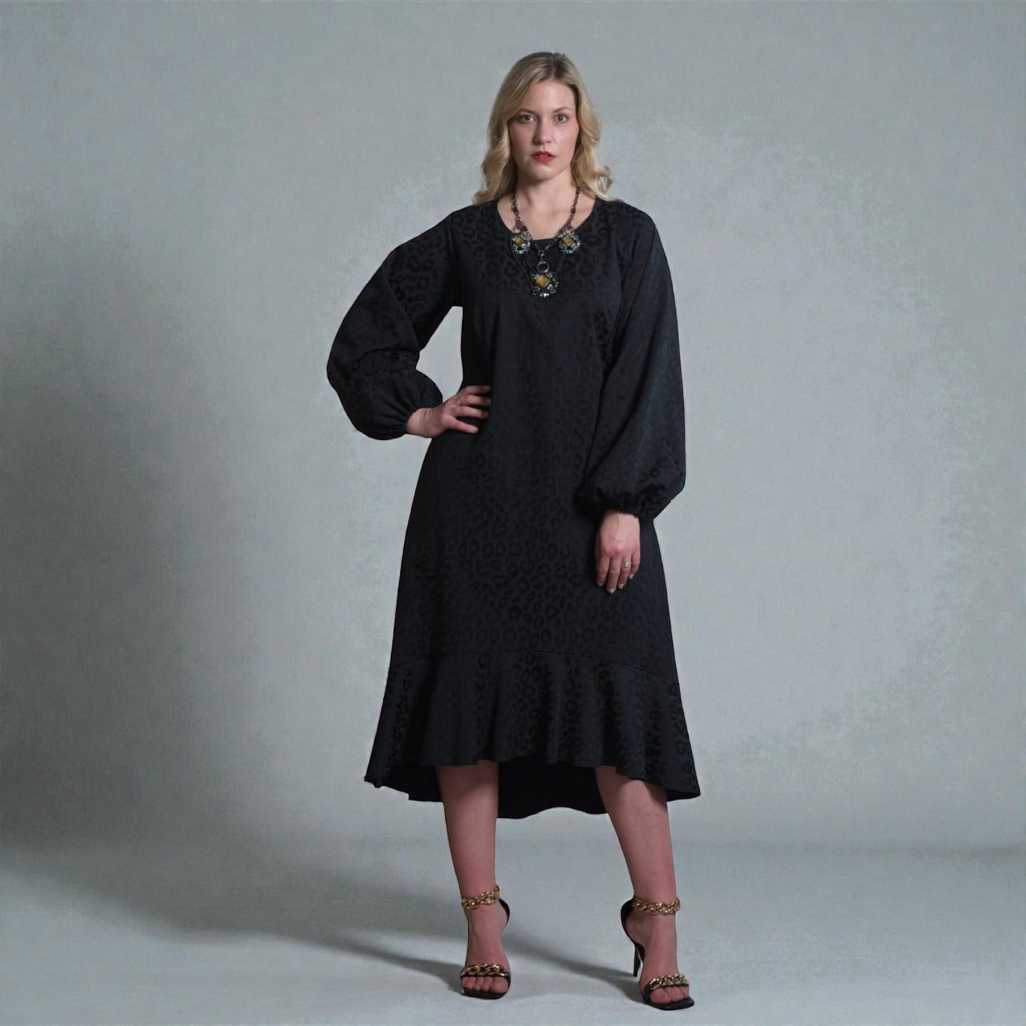 Women's Aphrodite Black Leopard Jacquard Dress styled with a gold necklace, black clutch and gold chain heels for a chic, coordinated look. Gray background.