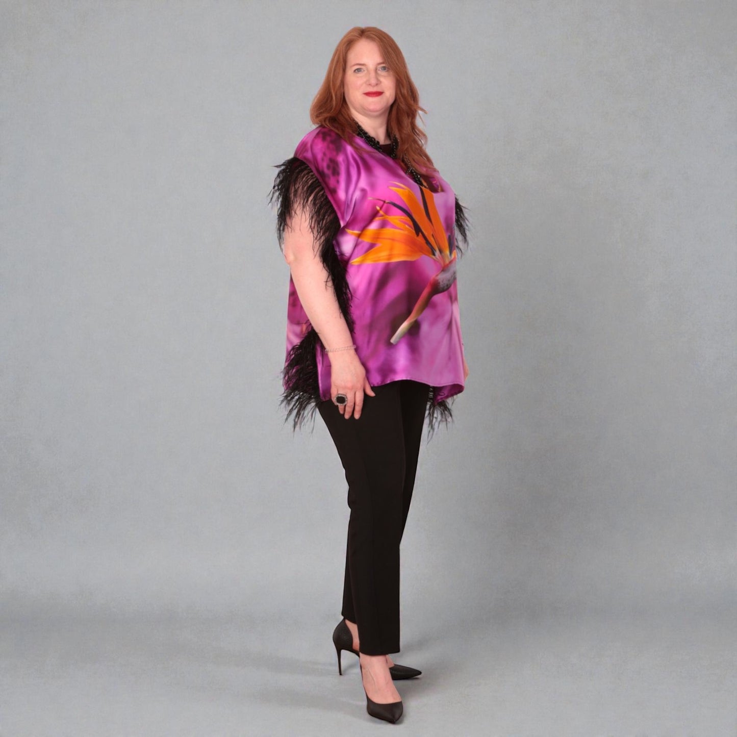 Women wearing a pink purple top with bird of paradise flower design and  black feather trim with black trousers on a plain background