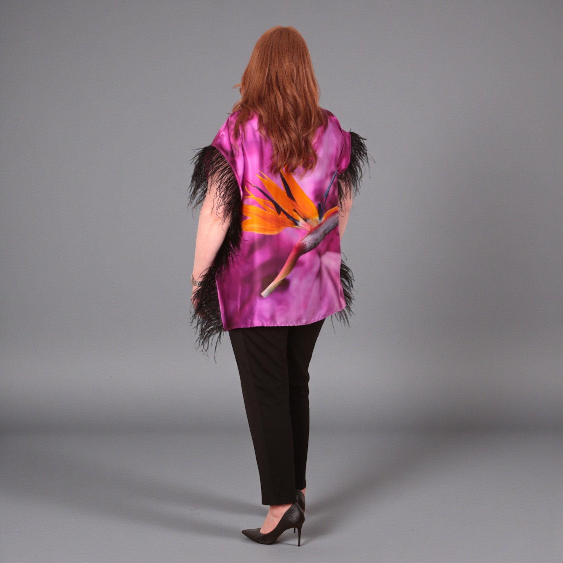 Women wearing a pink purple top with bird of paradise flower design and  black feather trim with black trousers on a plain background. Photographed from the back