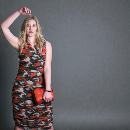Woman wearing a camouflage ruched midi dress with an orange clutch against a gray background