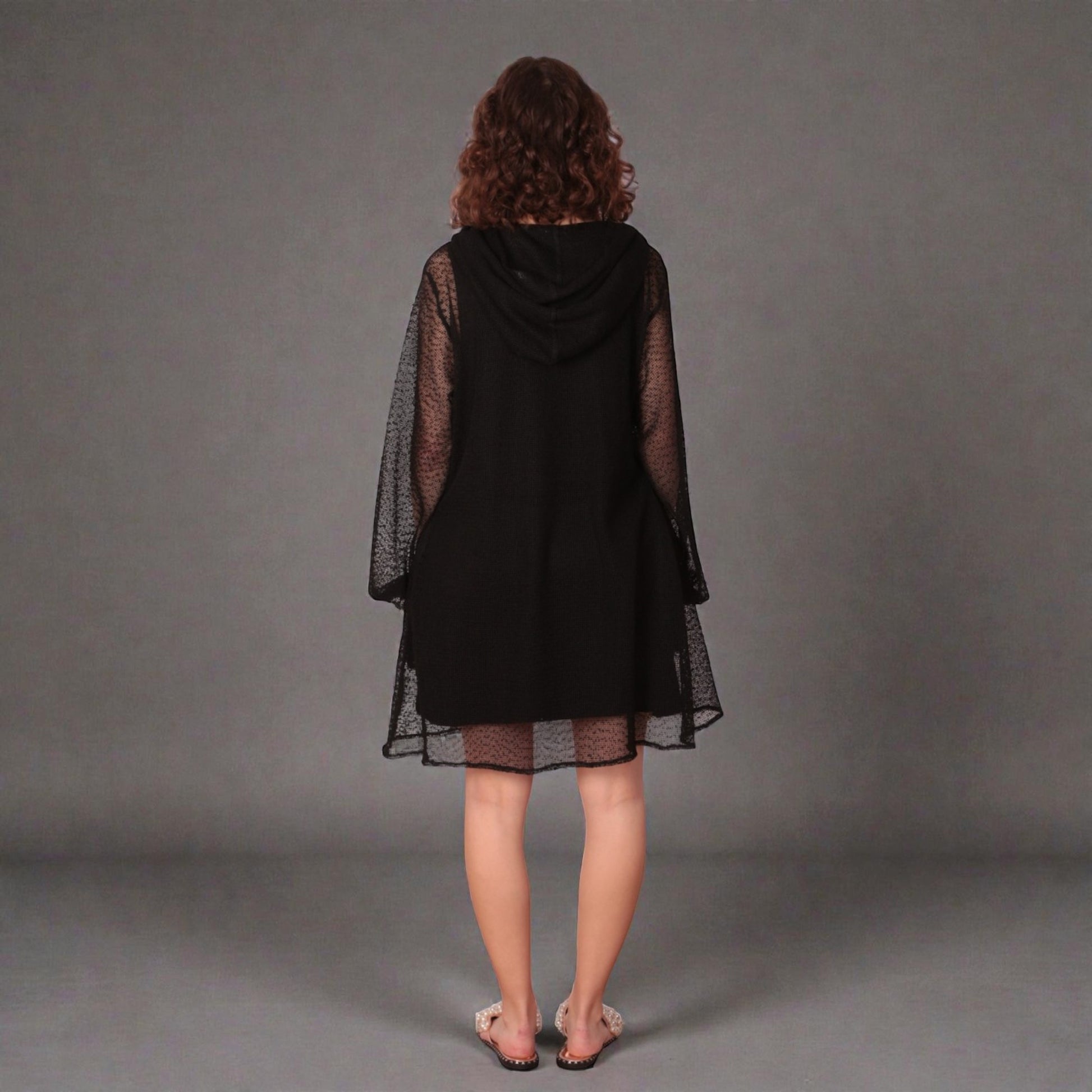 Person wearing a black dress with sheer sleeves, hoodie and black undergarment on a white background. photographed from the back