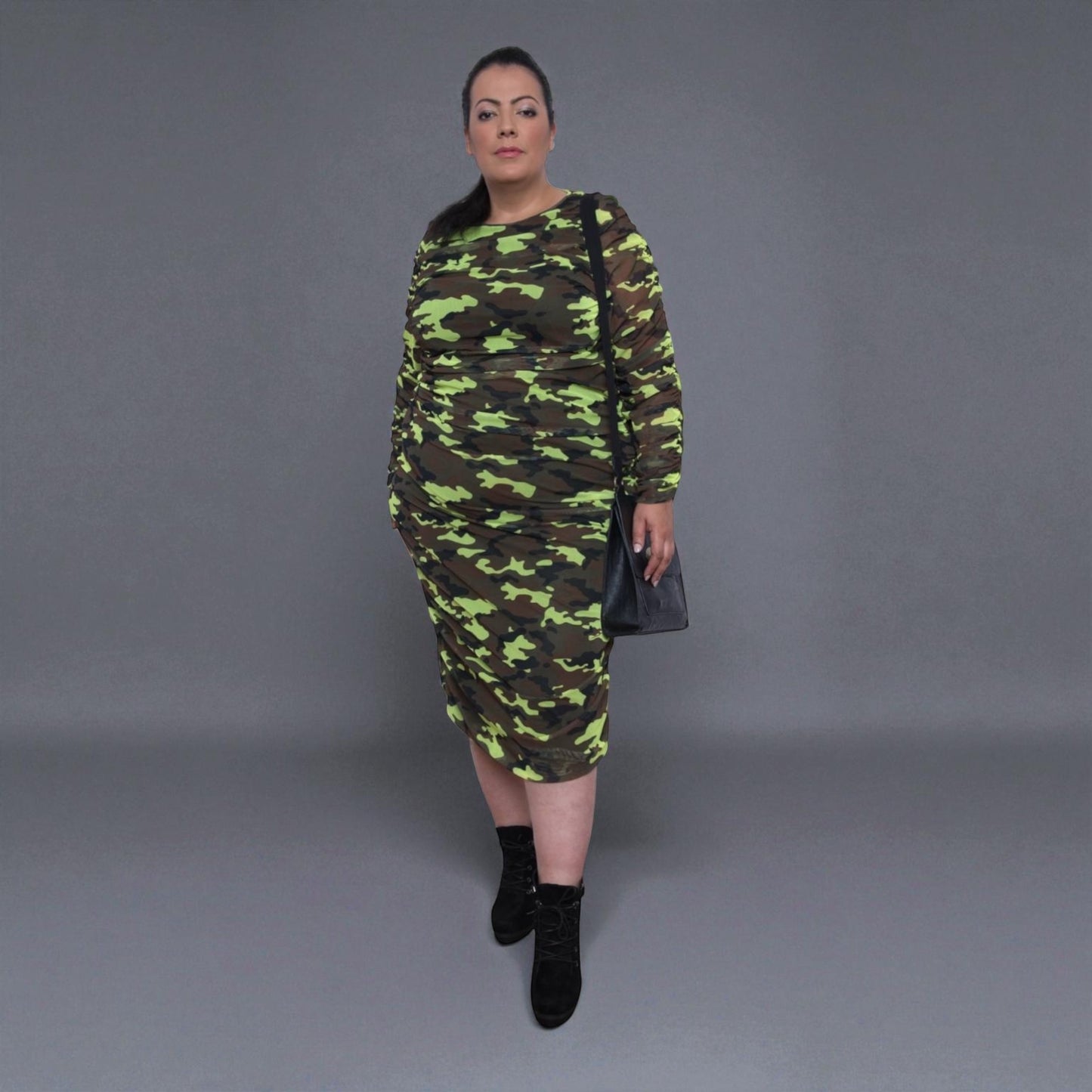 Plus Size Woman Neon Lime Brown Black Camouflage Bodycon Midi Dress styled with black boots for a bold and chic look. Grey Background.