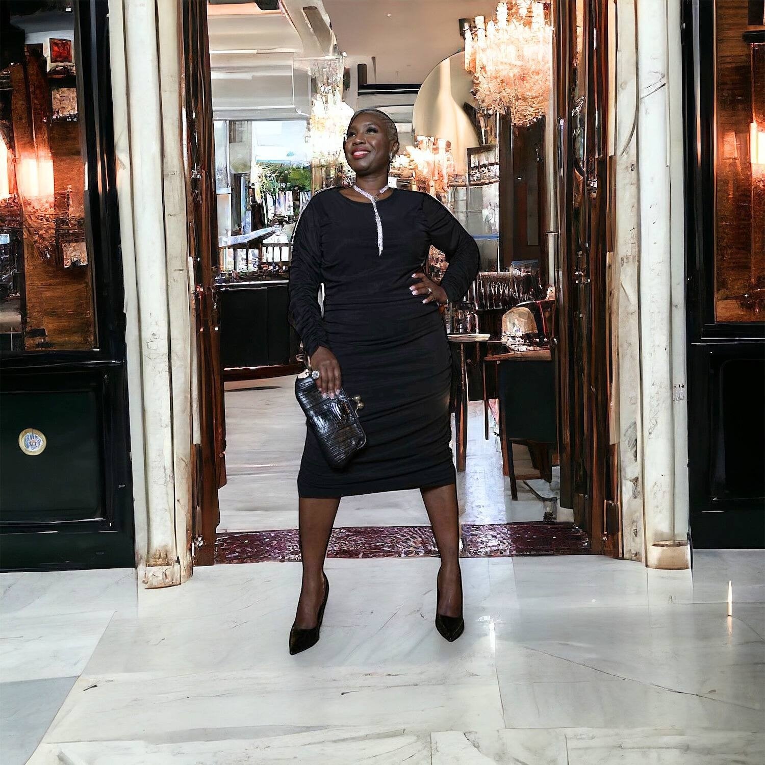 Plus-size woman wearing a black Boudicca Ruched Bodycon Midi Dress, styled with a sparkly necklace and black clutch, standing at the entrance of a restaurant.