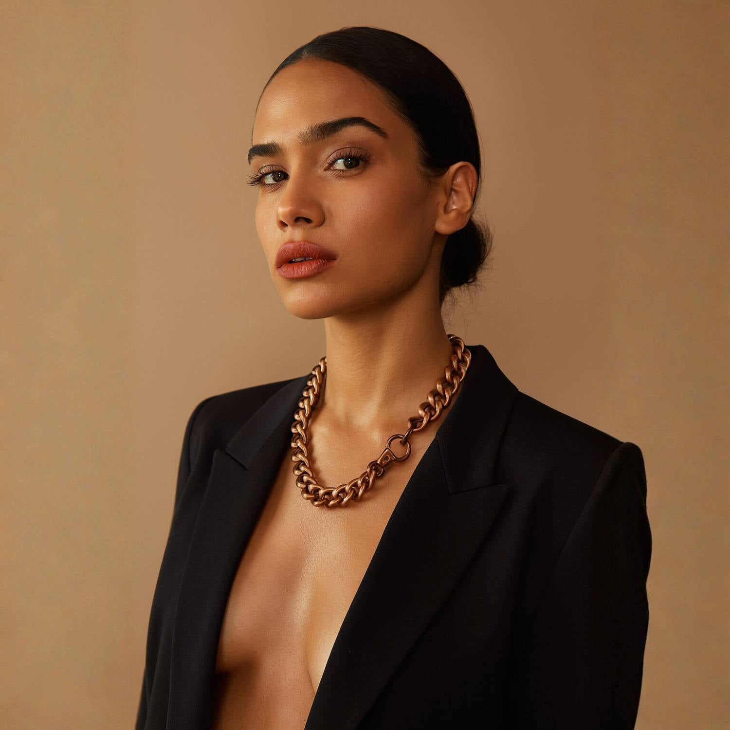 Woman wearing a black blazer and gold antique brass chain necklace against a beige background.