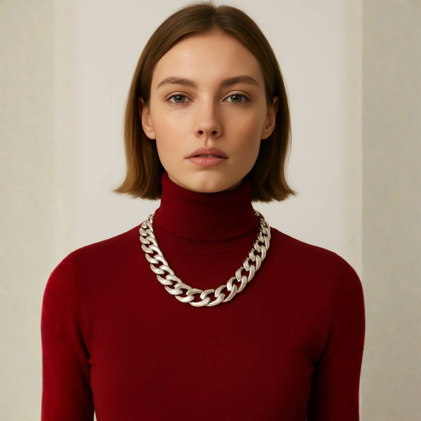 Woman wearing a red turtleneck sweater with a large silver chain necklace against a plain background