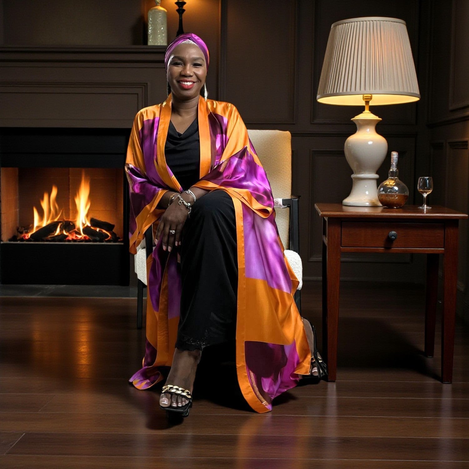 Woman sitting in a chair wearing a colorful  Bird of Paradise Pink Purple Silk Headscarf and  Kimono in a cozy room with a fireplace.