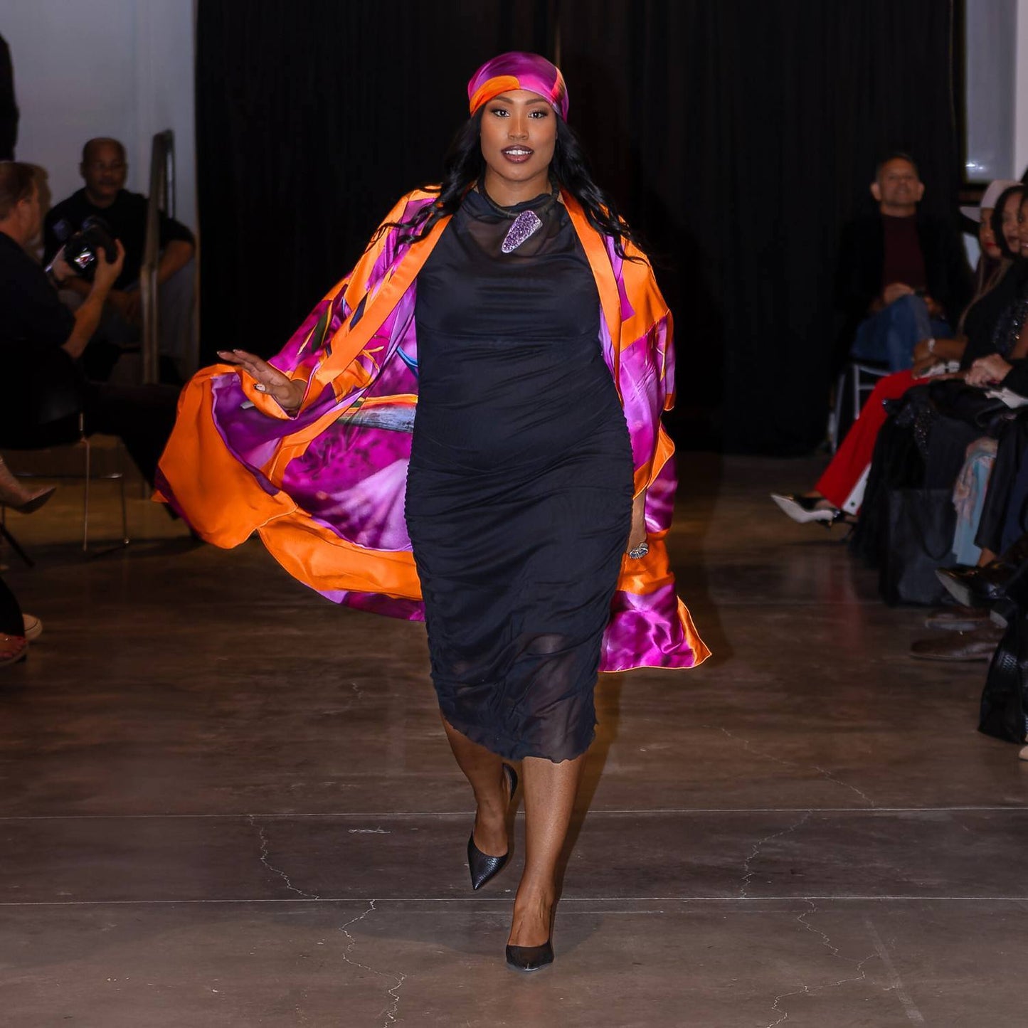 Model walking on a runway wearing a black Boudicca dress with a Bird of Paradise Pink Purple Silk Headscarf and Kimono.