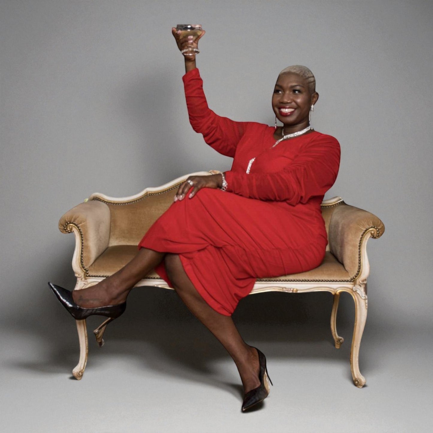 Woman in a Boudicca Tomato Red Ruched Midi Dress sitting on an ornate chair against a gray background