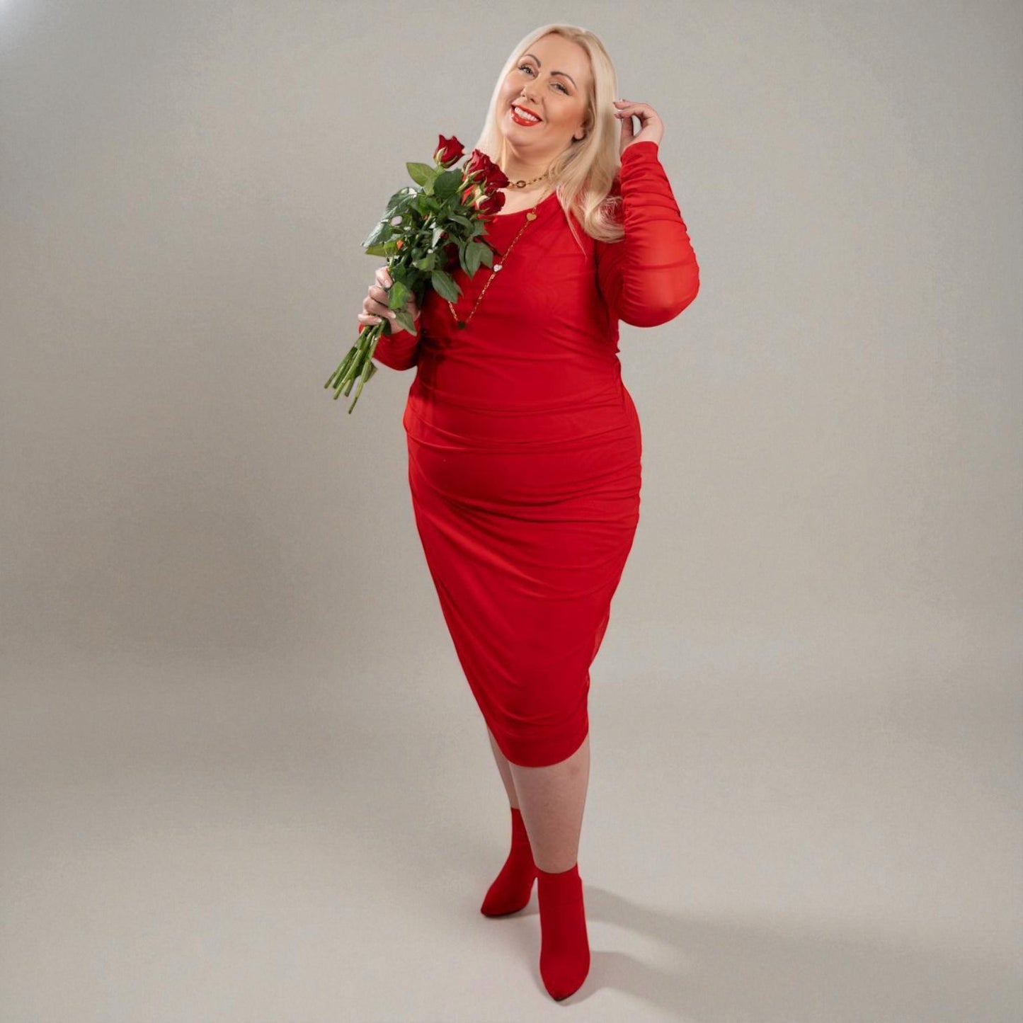 Woman in a Boudicca Tomato Red Ruched Midi Dress holding red roses flowers against a plain background