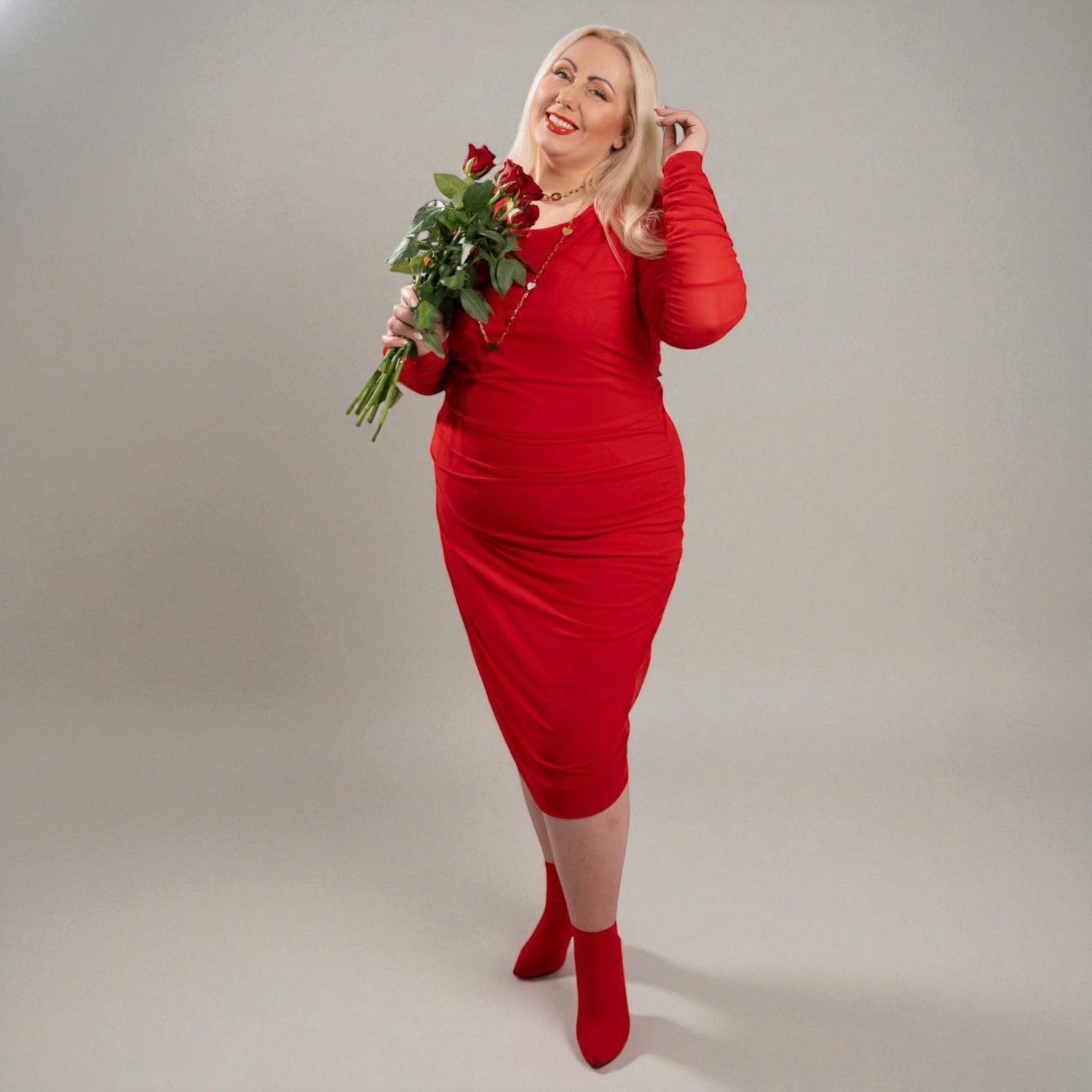 Woman in a Boudicca Tomato Red Ruched Midi Dress holding red roses flowers against a plain background