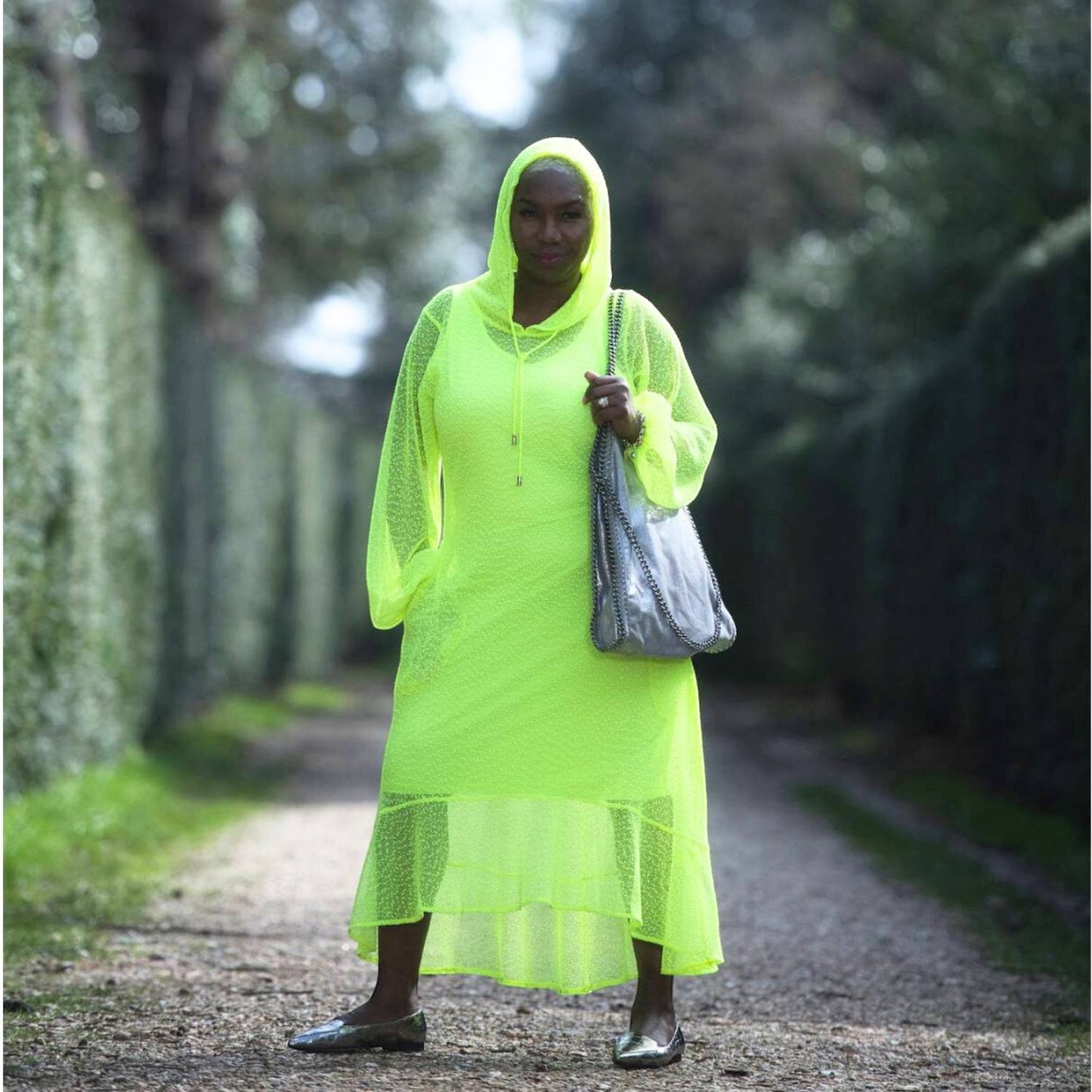 Sylvia Piechulla neon lime midi dress with hoodie, bishop sleeves, and flounce hem, worn by a model on a path.
